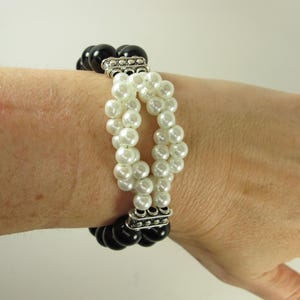 Puede incluir: Una pulsera de cuentas en blanco y negro con detalles plateados. La pulsera presenta grandes cuentas negras redondas en la parte superior e inferior, con una sección central de cuentas blancas con forma de perla. La pulsera se lleva en la muñeca.