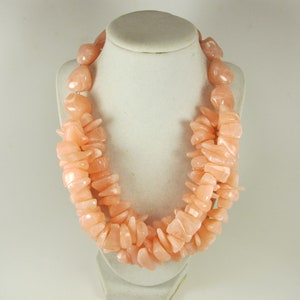 May include: A peach-coloured necklace displayed on a white bust. The necklace is composed of irregular, chunky, polished stones. The stones are strung together to form a double-strand necklace. The necklace is a statement piece.