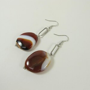 May include: A pair of dangle earrings featuring polished, oval-shaped stones in shades of brown, white, and black. The stones are connected to silver-colored metal hooks and rectangular links. The earrings are displayed against a white background.