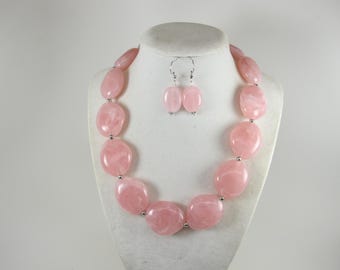 May include: A pink necklace and earring set. The necklace has large, oval beads in a soft pink colour, linked by silver-toned metal. The earrings match the necklace, creating a coordinated jewellery set. The beads are approximately 2.5 cm in length.