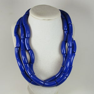 May include: A bold, cobalt blue necklace composed of multiple strands of cylindrical beads. The necklace is displayed on a white bust, showcasing its layered design and glossy finish. This statement piece is a striking accessory.