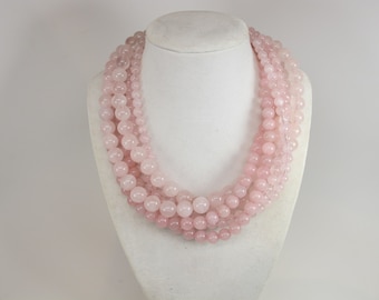 May include: A multi-strand necklace with rose quartz beads of different sizes. The necklace is displayed on a white bust, highlighting the soft pink colour and the varying bead sizes. A delicate and elegant jewellery piece.
