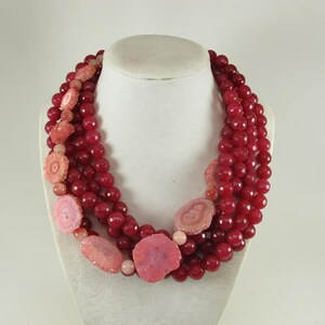 May include: A multi-strand necklace featuring deep red faceted beads interspersed with light pink, circular, textured stone slices. The necklace is arranged in several layers, creating a voluminous look with a mix of smooth and rough textures.