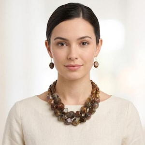 May include: A multi-strand necklace featuring polished, faceted brown and amber-toned beads, paired with matching drop earrings. The beads have a translucent, marbled appearance with varying shades of earthy brown, tan, and dark amber throughout.