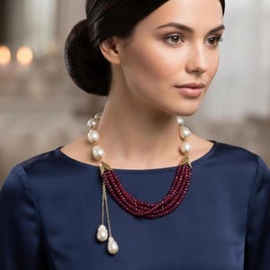 May include: A multi-strand necklace featuring deep red faceted beads arranged in a layered design. The necklace is accented with large, irregular white pearls and gold-toned metal leaf-shaped connectors, with two dangling pearl charms on a chain.