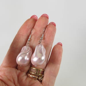 Puede incluir: Un par de pendientes de perlas rosas claras de estilo barroco con ganchos plateados. Los pendientes están sostenidos por una mano con esmalte de uñas rojo, mostrando el brillo nacarado de las perlas grandes de forma irregular.