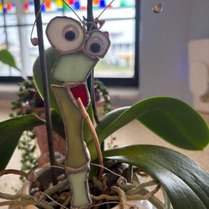 May include: A green stained glass bug with large white eyes and a red mouth is placed in a white pot with green plant roots. The bug has wire antennae.