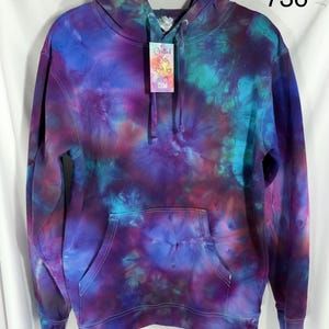 May include: A long-sleeve hooded sweatshirt featuring a tie-dye pattern in shades of purple, blue, and magenta. The hoodie includes a front kangaroo pocket, a drawstring hood, and a tag that reads Crafted With Love by Kim.