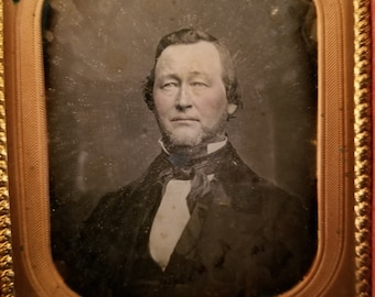 May include: A black and white portrait of a man in a dark suit and a white shirt with a bow tie. The portrait is framed in a gold frame with a red velvet mount.