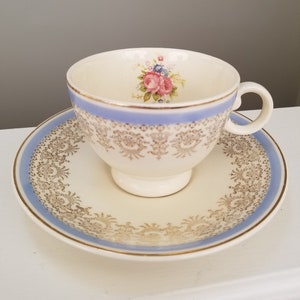 May include: A vintage teacup and saucer set with a floral design. The cup and saucer are white with a blue and gold trim. The floral design features pink, blue, and green flowers.