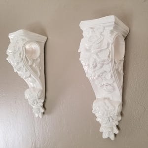 May include: Two white decorative wall corbels with ornate floral and scrollwork designs. The corbels are mounted on a neutral-colored wall, showcasing their detailed relief patterns. These architectural elements add a touch of elegance and are suitable for supporting shelves or decorative items.