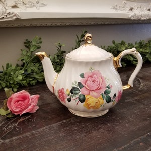 May include: White teapot with a gold trim and floral design featuring pink and yellow roses. The teapot is sitting on a wooden surface.
