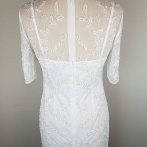 May include: White beaded dress with a sheer bodice and a fitted silhouette. The dress has a back zipper closure and a delicate floral pattern.