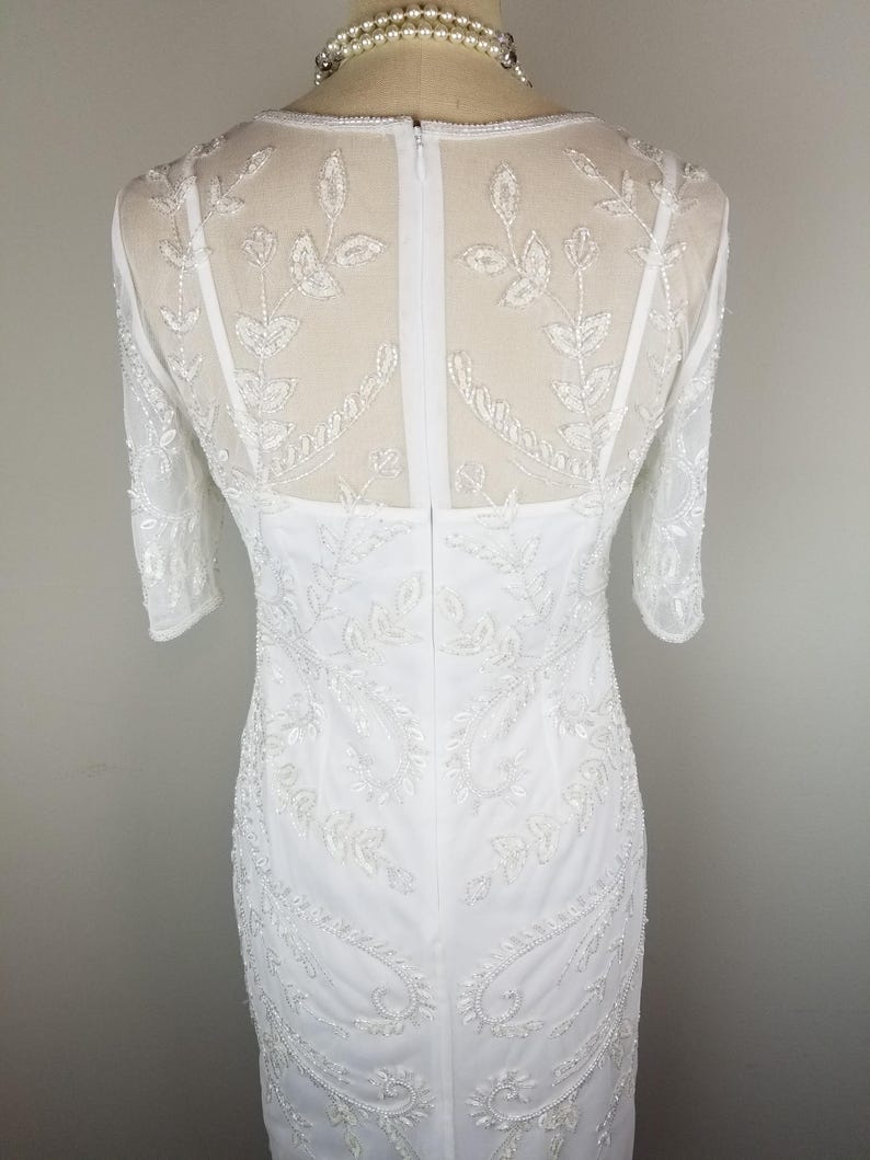 May include: White beaded dress with a sheer bodice and a fitted silhouette. The dress has a back zipper closure and a delicate floral pattern.