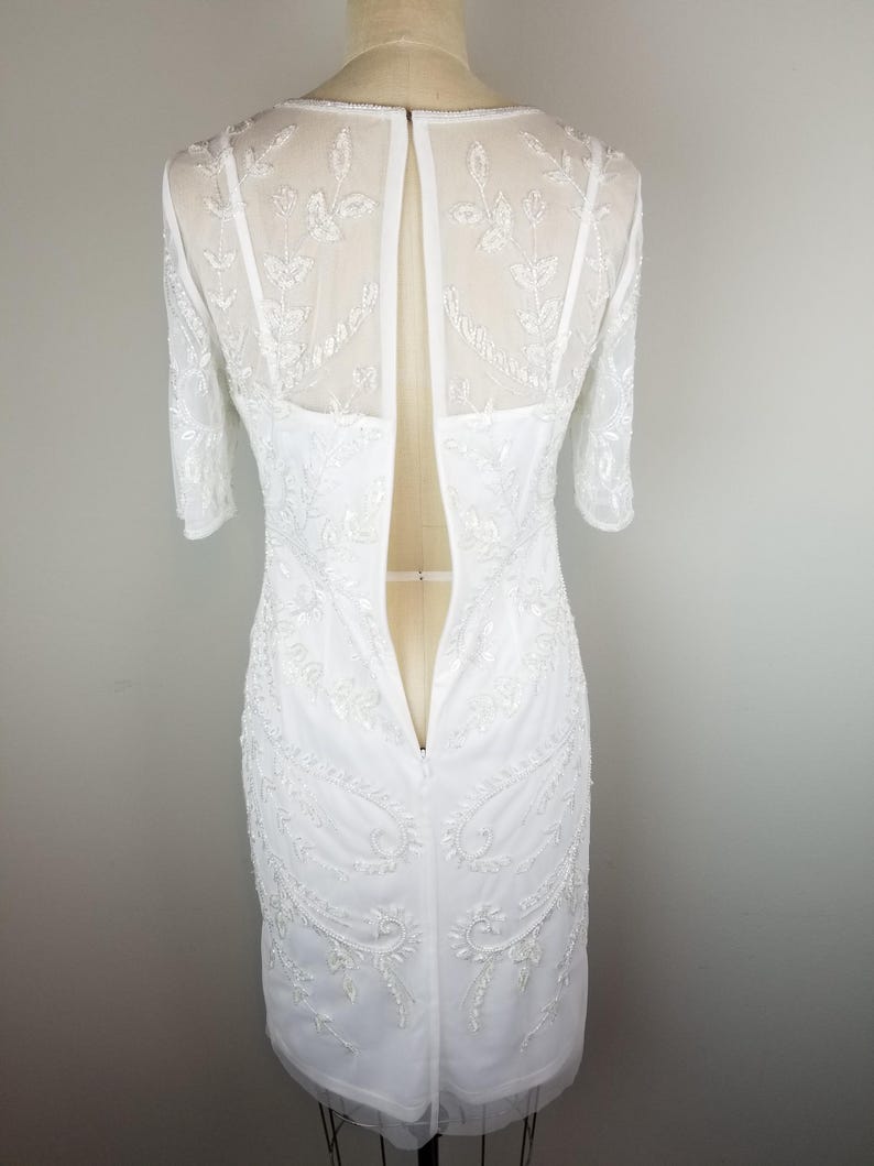 May include: White beaded dress with a sheer back and a zipper closure. The dress has a fitted silhouette and a short sleeve.