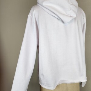 Puede incluir: Una sudadera blanca de manga larga con capucha, corte holgado, capucha con escote fruncido y dobladillo sin rematar. La prenda se muestra desde la parte posterior, exhibiendo el tejido blanco liso y la estructura de la capucha.