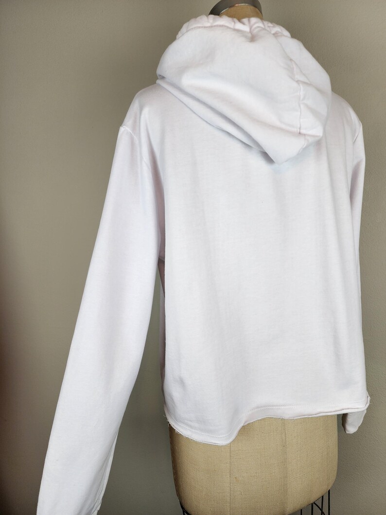Puede incluir: Una sudadera blanca de manga larga con capucha, corte holgado, capucha con escote fruncido y dobladillo sin rematar. La prenda se muestra desde la parte posterior, exhibiendo el tejido blanco liso y la estructura de la capucha.
