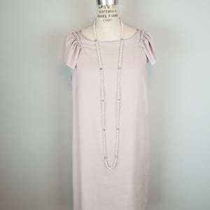 Può includere: Abito malva chiaro con maniche corte e scollatura rotonda, esposto su un manichino vintage. L'abito presenta finiture argentate sulle spalle e una lunga collana di perle. Design semplice ed elegante.