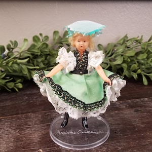 Puede incluir: Una muñeca de porcelana que lleva un vestido verde y blanco con ribetes de encaje y un sombrero azul. La muñeca está de pie sobre una base de plástico transparente con el texto "Made in Austria" impreso en ella.