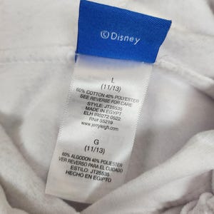 Puede incluir: Una prenda blanca con una etiqueta de marca azul de Disney y una etiqueta de cuidado blanca. La etiqueta muestra la talla L (11/13), composici&oacute;n de 60% algod&oacute;n y 40% poli&eacute;ster, estilo JT25535, origen Egipto y la direcci&oacute;n web jerryleigh.com.