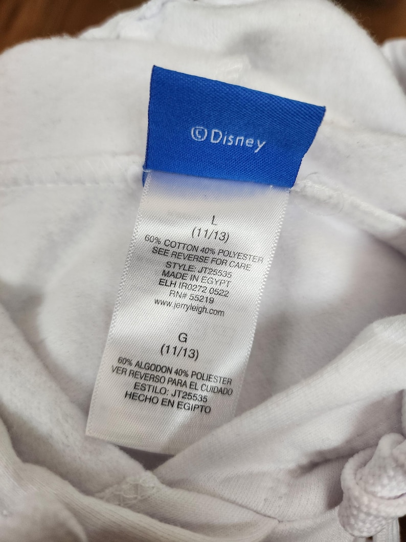 Puede incluir: Una prenda blanca con una etiqueta de marca azul de Disney y una etiqueta de cuidado blanca. La etiqueta muestra la talla L (11/13), composici&oacute;n de 60% algod&oacute;n y 40% poli&eacute;ster, estilo JT25535, origen Egipto y la direcci&oacute;n web jerryleigh.com.