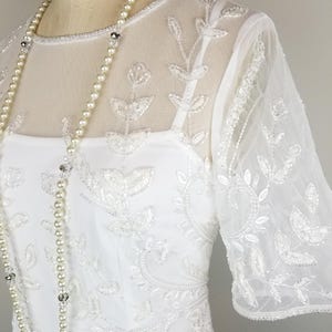 May include: A white beaded dress with a sheer overlay and a pearl necklace. The dress has a round neckline and short sleeves. The beading is in a floral pattern.