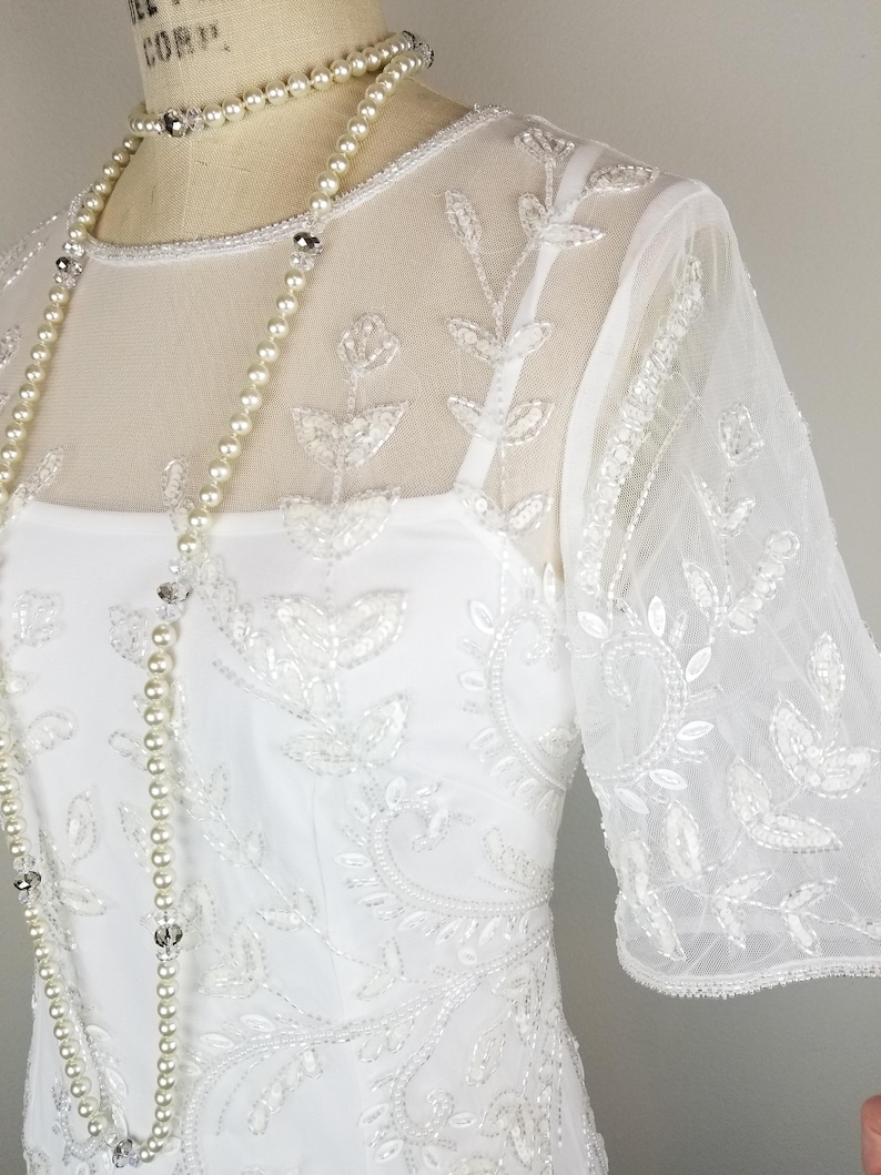 May include: A white beaded dress with a sheer overlay and a pearl necklace. The dress has a round neckline and short sleeves. The beading is in a floral pattern.