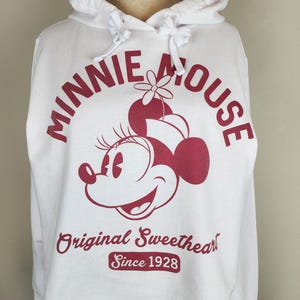 Puede incluir: Una sudadera con capucha blanca que presenta un gr&aacute;fico granate de Minnie Mouse con un sombrero con una flor. El texto dice Minnie Mouse, Original Sweetheart y Since 1928 en fuente granate. La sudadera incluye una capucha con cord&oacute;n y mangas largas.
