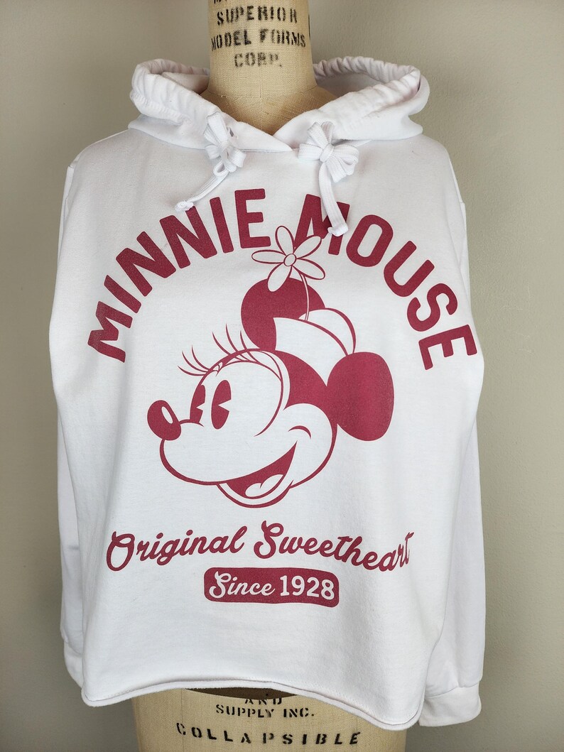 Puede incluir: Una sudadera con capucha blanca que presenta un gr&aacute;fico granate de Minnie Mouse con un sombrero con una flor. El texto dice Minnie Mouse, Original Sweetheart y Since 1928 en fuente granate. La sudadera incluye una capucha con cord&oacute;n y mangas largas.