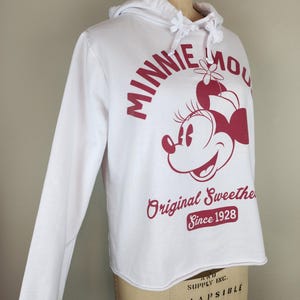 Puede incluir: Una sudadera blanca de manga larga con capucha y un gr&aacute;fico rojo de Minnie Mouse. El texto dice MINNIE MOUSE Original Sweetheart Since 1928. La sudadera tiene un cord&oacute;n en el cuello y un borde inferior sin dobladillo, sobre un maniqu&iacute;.