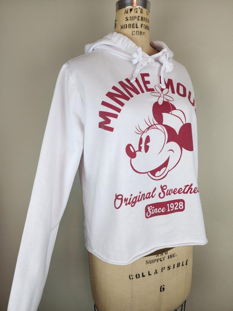 Puede incluir: Una sudadera blanca de manga larga con capucha y un gr&aacute;fico rojo de Minnie Mouse. El texto dice MINNIE MOUSE Original Sweetheart Since 1928. La sudadera tiene un cord&oacute;n en el cuello y un borde inferior sin dobladillo, sobre un maniqu&iacute;.