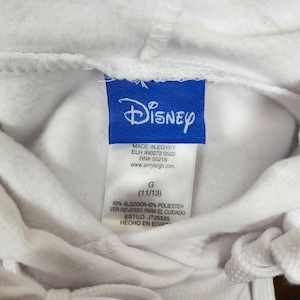 Puede incluir: Una sudadera con capucha blanca con una etiqueta de marca Disney azul en el cuello. La etiqueta incluye el texto Made in Egypt, RN 55219, www.jerryleigh.com, talla G (11/13), 60% algodon 40% poliester, ver reverso para el cuidado y estilo JT25535.