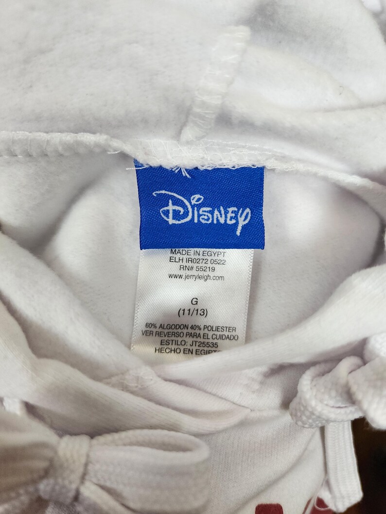 Puede incluir: Una sudadera con capucha blanca con una etiqueta de marca Disney azul en el cuello. La etiqueta incluye el texto Made in Egypt, RN 55219, www.jerryleigh.com, talla G (11/13), 60% algodon 40% poliester, ver reverso para el cuidado y estilo JT25535.