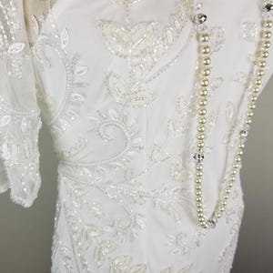 May include: A white beaded dress with a sheer overlay and a pearl necklace. The dress features a floral design with delicate beading.