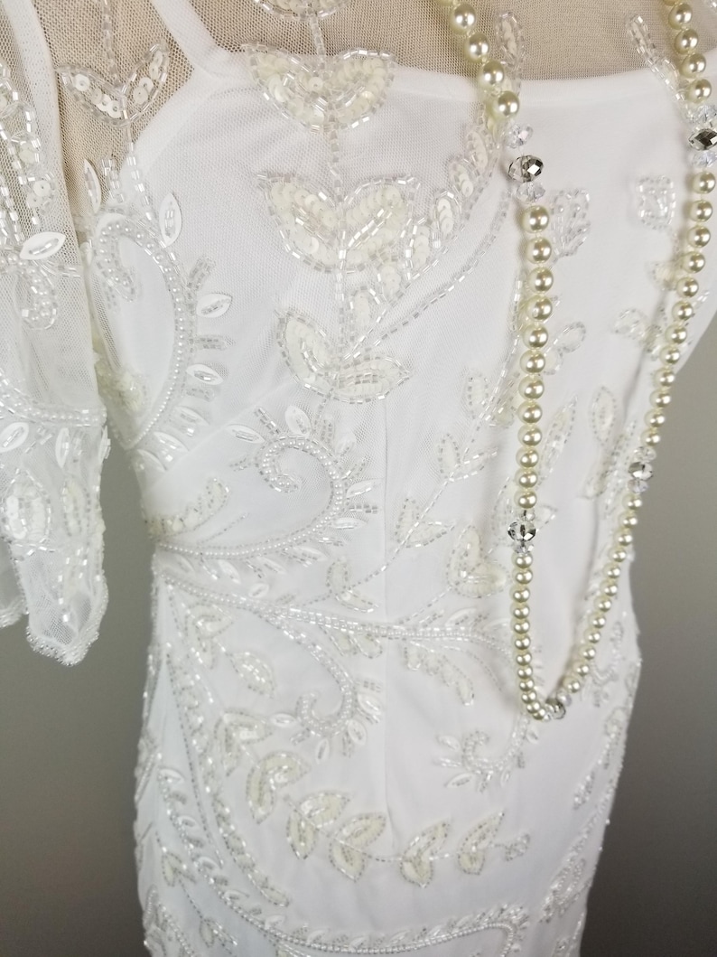 May include: A white beaded dress with a sheer overlay and a pearl necklace. The dress features a floral design with delicate beading.