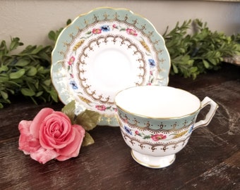 May include: A vintage teacup and saucer set with a light blue border and gold trim. The cup and saucer feature a floral design with pink, blue, and yellow flowers. A pink rose is placed next to the teacup.