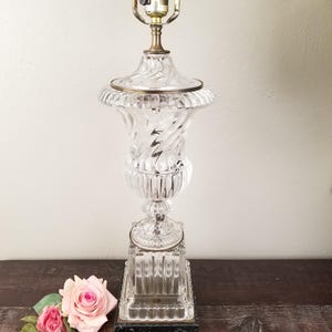 May include: A clear glass table lamp with a decorative urn-shaped body and a brass-colored lamp holder. The lamp has a spiral design and sits on a rectangular base. Pink roses are on the table next to the lamp.