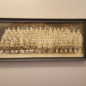 Puede incluir: Una fotografía panorámica en tono sepia de un gran grupo de soldados con uniforme, dispuestos en varias filas. La imagen está dentro de un marco rectangular oscuro y texturizado. Se observa texto tenue en el borde inferior de la foto.