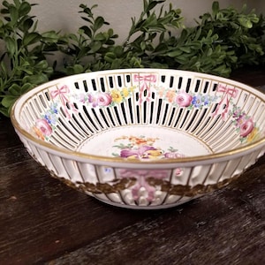 Puede incluir: Cesta de porcelana blanca con diseño calado, borde dorado y detalles florales y de lazo. El interior tiene un arreglo floral central. Una pieza decorativa para servir o exhibir.