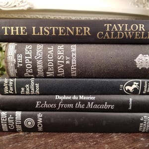 May include: A stack of five black hardcover books with white text on the spines. The top book reads "The Listener Taylor Caldwell". The second book reads "The People's Common Sense Medical Adviser by RV. Pierce MD". The third book reads "The Black Stallion's Ghost Walter Farley". The fourth book reads "Echoes from the Macabre Daphne du Maurier". The bottom book reads "The Amateur Gun-Craftsman Howe".
