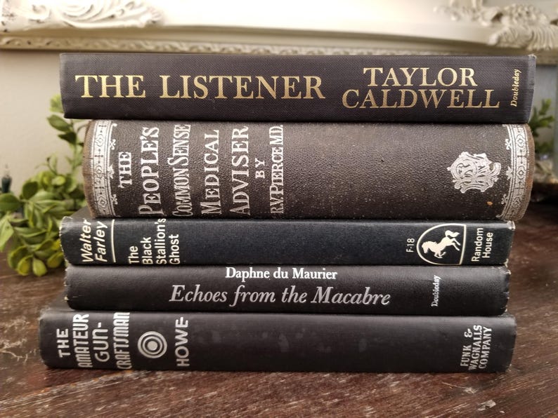 May include: A stack of five black hardcover books with white text on the spines. The top book reads "The Listener Taylor Caldwell". The second book reads "The People's Common Sense Medical Adviser by RV. Pierce MD". The third book reads "The Black Stallion's Ghost Walter Farley". The fourth book reads "Echoes from the Macabre Daphne du Maurier". The bottom book reads "The Amateur Gun-Craftsman Howe".