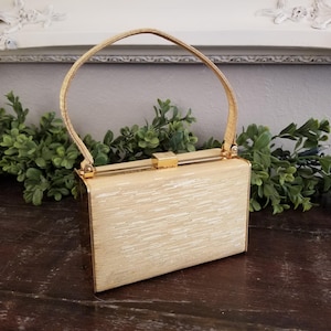May include: A vintage rectangular handbag with a gold-toned handle and clasp. The bag's body has a textured, light beige appearance. The handle is arched, and the clasp is rectangular. The bag is sitting on a dark wooden surface.