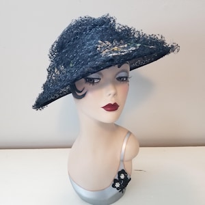 May include: A dark blue hat with a wide brim and a delicate, lacy texture. The hat is decorated with subtle floral accents. The hat is displayed on a mannequin head with a painted face and red lipstick.