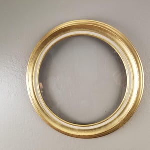 May include: A round, gold-colored picture frame with a white inner border. The frame is set against a light gray background. The frame is likely for displaying photos or artwork.