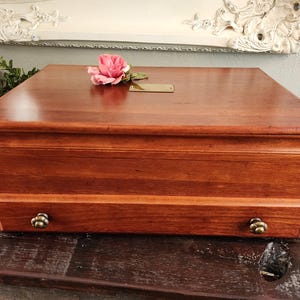May include: A rectangular wooden box with a reddish-brown finish, featuring a hinged lid, a single front drawer with two small brass-toned floral knobs, and a small rectangular brass-toned metal plate centred on the top surface.