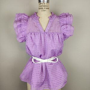 May include: A lavender blouse featuring a checkered pattern, a V-neckline with a ruffled collar, and short ruffled sleeves. The garment is cinched at the waist with a thin white belt tied in a bow, creating a peplum silhouette on the torso.