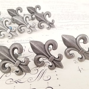 Pode incluir: Seis puxadores de gaveta de metal prateado em forma de flor-de-lis com um design decorativo.