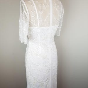 May include: A white beaded dress with a sheer bodice and short sleeves. The dress has a fitted silhouette and a delicate floral pattern.