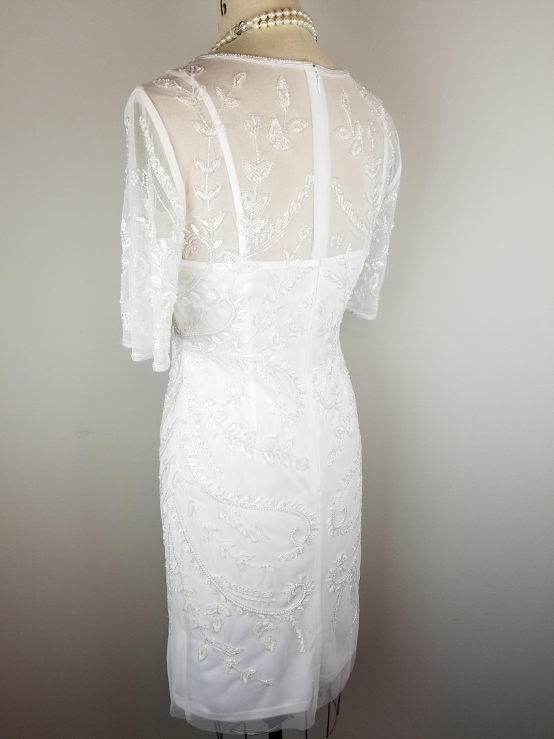 May include: A white beaded dress with a sheer bodice and short sleeves. The dress has a fitted silhouette and a delicate floral pattern.