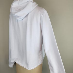 Puede incluir: Una sudadera blanca lisa de manga larga con capucha sin cordones, mangas largas con pu&ntilde;os acanalados y un dobladillo recto. La prenda se muestra sobre un maniqu&iacute;, exhibiendo la vista posterior de la tela blanca s&oacute;lida.